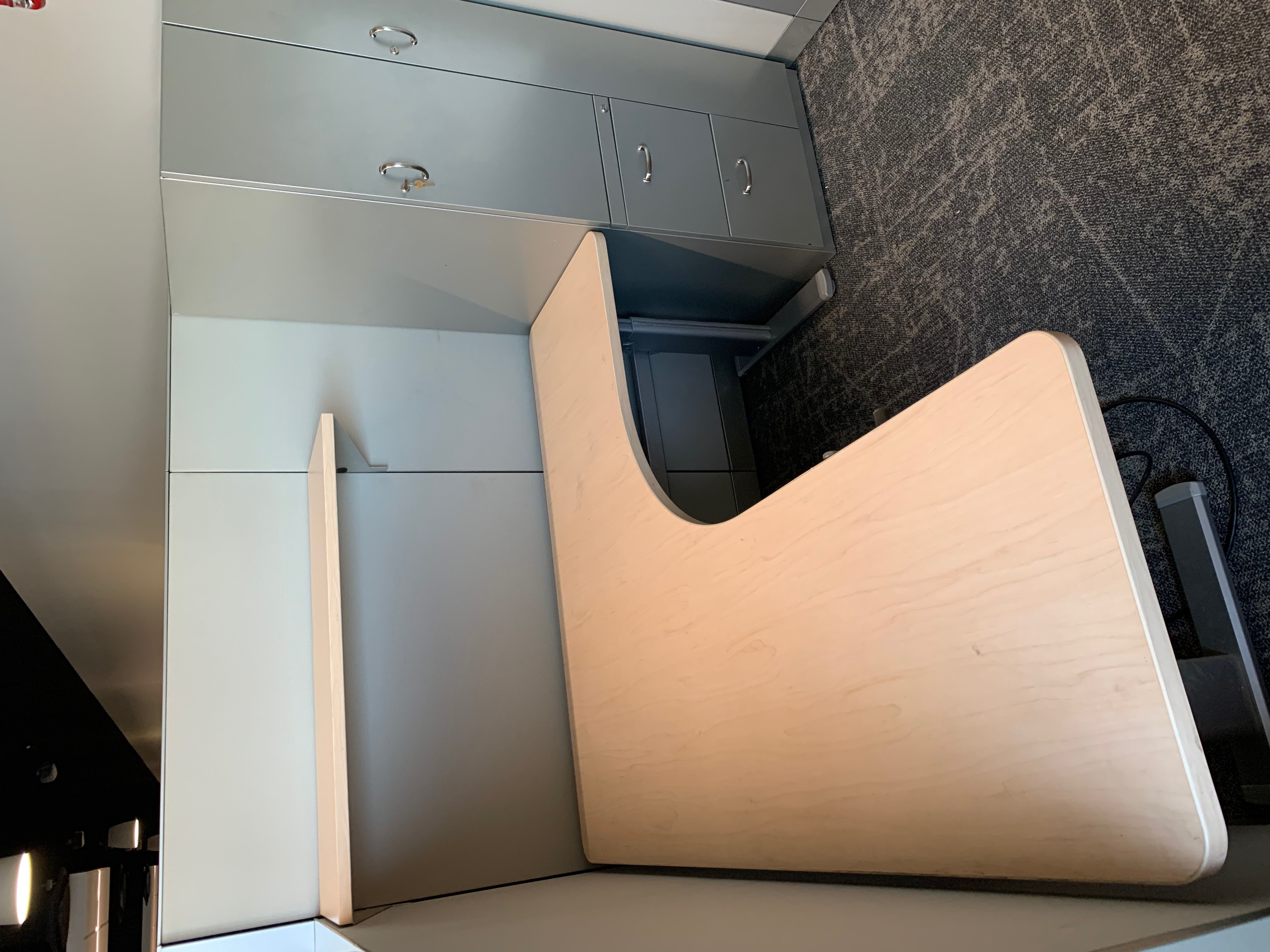 Cubicle workstation with an L-shaped maple desk, overhead shelf, and a storage tower